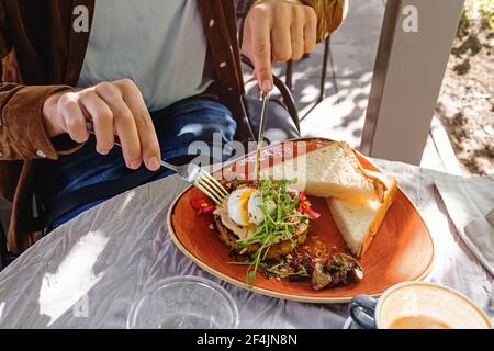 Männliche Hände schneiden pochiertes Ei mit Gabel und Messer. Stockfoto