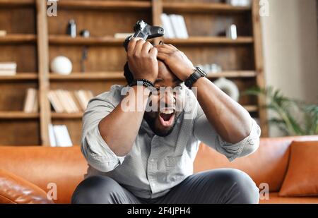Vorderansicht Porträt des jungen afroamerikanischen Kerl auf der Couch im Wohnzimmer sitzen, halten Kopf in den Händen und Joystick, schreien, weil Videospiel-Spiel verloren, männlich süchtig Gamer Gefühl verrückt Stockfoto