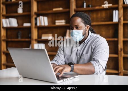 Konzentrierter junger afroamerikanischer Student in schützender Gesichtsmaske, sitzt im Büro mit Bücherregalen oder Bibliothek und tippen auf Laptop, arbeiten an Projekt-, Sicherheits-, Schutz- und Präventionskonzepten Stockfoto