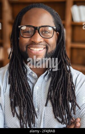 Vertikales Porträt von lächelndem jungen gutaussehenden afroamerikanischen Mann mit Dreadlocks und Brille stehend mit Armen gefaltet und Blick auf die Kamera, optimistischen schwarzen Mann Büroleiter ist bereit zu arbeiten Stockfoto