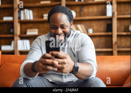 Glücklich lachend gemischter Mann auf der Couch sitzen, zu Hause bleiben, SMS-Chat mit Freunden auf dem Handy, mit Dating-App, flirten, Comedy-Show ansehen, lustiges Video, Online-Spiele spielen Stockfoto