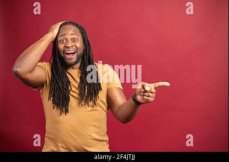 Erstaunt afroamerikanischen Mann mit Dreadlocks stehen isoliert über rotem Hintergrund halten Kopf, zeigt mit dem Finger auf den leeren Raum für Werbung, Mockup Bild, überprüfen Sie dies, lustiges Konzept Stockfoto