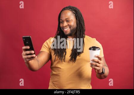 Porträt eines afroamerikanischen Blogger mit Dreadlocks isoliert auf der roten Wand hält Kaffee Tasse, Streaming-Video online über Handy, Selfie für Social Media-Netzwerk, posieren, lächeln Stockfoto