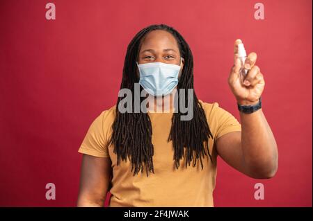 Junger afroamerikanischer Mann mit Dreadlocks tragen Schutzmaske isoliert auf rotem Hintergrund stehend und Sprühen halten Desinfektionsmittel vor der Arbeit, Hygiene und Sicherheitskonzept während covid-19 Stockfoto