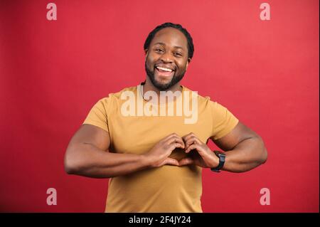 Romantik-Konzept. Lächelnd und gut aussehend dunkelhäutig Mann zeigt Herzform mit Händen, glücklich afrikanischen Kerl macht Liebe Geste, isoliert auf rot Stockfoto