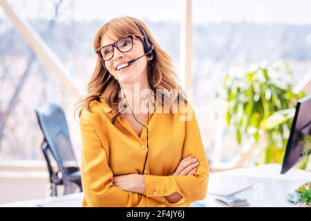 Eine reife Geschäftsfrau im Headset spricht per Telefonkonferenz, während sie am Schreibtisch steht. Heimbüro. Stockfoto