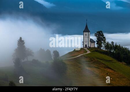 Jamnik, Slowenien - Foggy Sommermorgen an Jamnik St.Primoz Bergkirche. Der Nebel geht sanft um die kleine Hügelkapelle mit grünen Feldern und Stockfoto