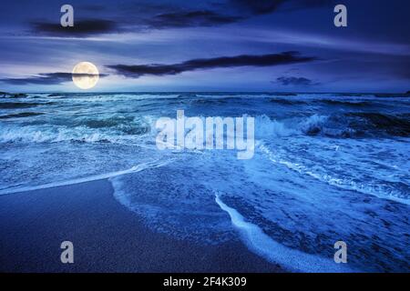 Meeresflut bei bewölktem Sonnenaufgang. Grüne Wellen krachen goldenem Sandstrand im Vollmondlicht. Sturmwetter nähert sich. Sommerferienkonzept Stockfoto