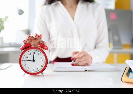 Geschäftsfrau an ihrem Schreibtisch hält ihre Hand auf rotem Alarm Uhr und nimmt Einträge in Tagebuch Stockfoto