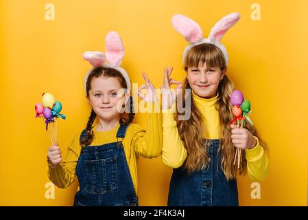 Portrait von niedlichen schönen kleinen Schwestern Mädchen in rosa Hase flauschige Ohren halten kleine Ostern farbige Eier, macht okay Geste, sagt ok Stockfoto