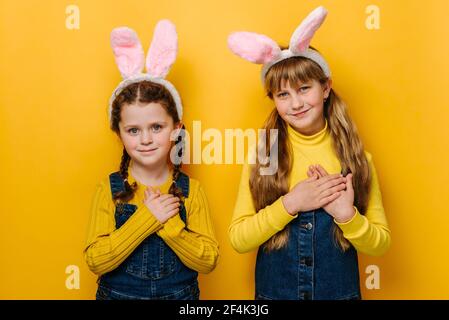 Portrait von zwei glücklich lächelnden Mädchen Kind in rosa Hase flauschige Ohren legte gefaltete Palmen auf Herz, isoliert über gelben Studio-Hintergrund. Konzept ostern Stockfoto