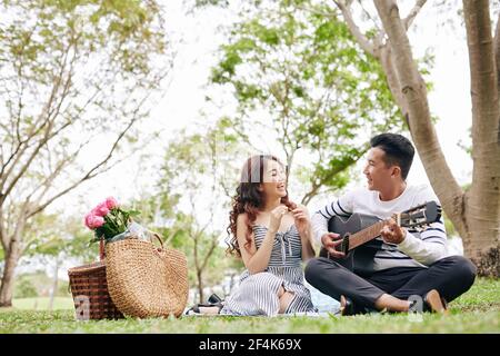 Schöner junger Vietnamesischer Mann, der Gitarre spielt und Lied singt Freundin, wenn sie mit Picknick im Park Stockfoto