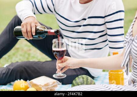 Beschnittenes Bild eines jungen Mannes, der Rotwein in Glas gießt Von Freundin, wenn sie Picknick im Park genießen Stockfoto