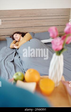 Schöne junge Frau schlafen im Bett, wenn Mann bringt sie Tablett mit Frühstück und Blumen Stockfoto