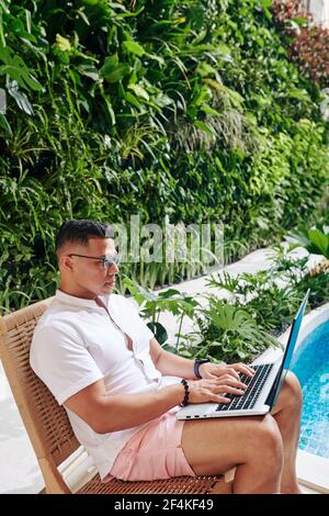 Passen gut aussehende seriöse Geschäftsmann in einer Brille sitzen auf einem Stuhl am Pool und arbeiten auf Laptop, beantworten E-Mails und Dokumente zu überprüfen Stockfoto