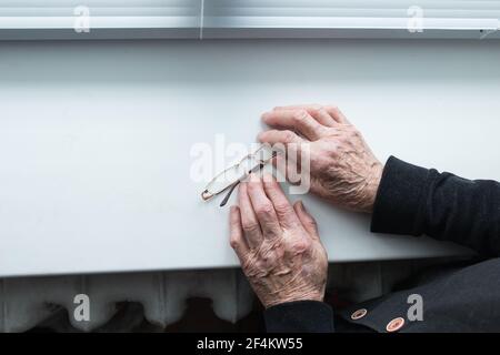 Ältere Menschen, die unter Einsamkeit leiden. Die Hände einer älteren Frau halten eine Brille auf der Fensterbank. Stockfoto