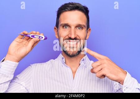 Junger schöner Mann Kontrolle Temperatur halten Thermometer über lila Hintergrund Lächelnd glücklich zeigen mit Hand und Finger Stockfoto