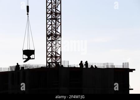 Silhouetten von Arbeitern und Turmdrehkran mit Ladung auf der Baustelle gegen den Himmel. Wohnungsbau, Bauherren arbeiten auf Gerüsten Stockfoto