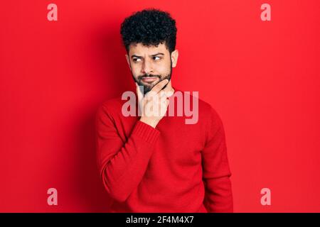 Junger arabischer Mann mit Bart trägt lässigen roten Pullover mit Hand auf Kinn Denken über Frage, nachdenklichen Ausdruck. Lächelnd mit nachdenklichen Gesicht. doub Stockfoto