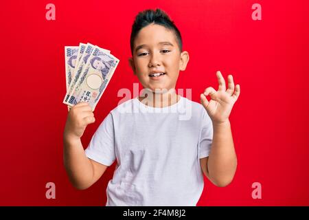 Kleiner Junge hispanic Kind hält 5000 japanische Yen Banknoten tun ok Zeichen mit Fingern, lächelnd freundlich gestikulieren ausgezeichnetes Symbol Stockfoto