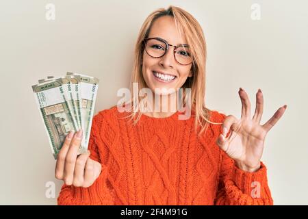 Schöne blonde Frau hält indische Rupie Banknoten tun ok Zeichen mit Fingern, lächelnd freundlich gestikulieren ausgezeichnete Symbol Stockfoto