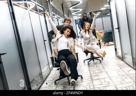 Lustige junge Erwachsene Geschäftsleute machen ein Rennen auf Stühlen im modernen Büro. Multirassisch freundliche Kollegen verbringen zusammen ihre Arbeitspause, Ruhe von Brainstorming Stockfoto