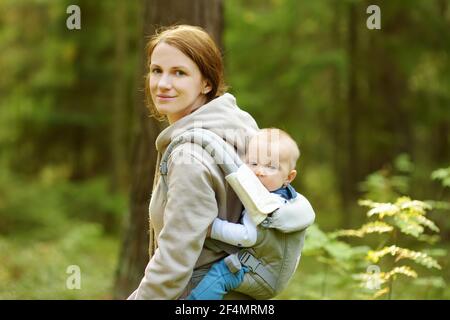 Junge Mutter mit ihrem kleinen Sohn in einer Babytrage Spaß beim Wandern in einem Wald an schönen Sommertag. Neue Mutter, die einen Spaziergang mit ihrem kleinen Sohn. Auto Stockfoto