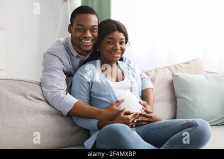 Lächelnder schwarzer Mann umarmt seine schwangere Frau, die auf dem Sofa sitzt Stockfoto