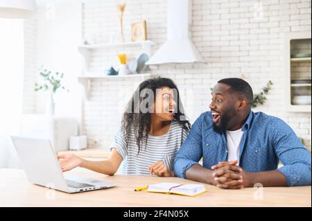 Aufgeregt multi ethnischen Paar in der Liebe nicht glauben, ihre Augen, sitzt am Küchentisch vor einem Laptop, schaut sich gegenseitig mit Schock, erhielt Hypothek Genehmigung per Post, gewinnen im Lotto Stockfoto