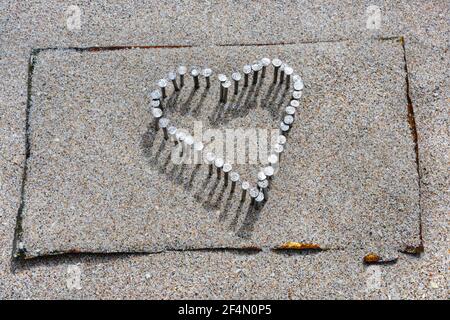 Herz aus Nägeln in abgeflachtem Zinn über Holzbrett gehämmert, mit Strandsand bedeckt. Stockfoto