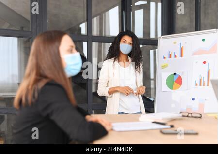 Multiethnische Arbeitsgruppe Brainstorming im zeitgenössischen Büro während der Pandemie, eine vielfältige Kollegen tragen medizinische Maske innen. Eine afroamerikanische Frau mit bedecktem Gesicht in der Nähe des Whiteboards Stockfoto