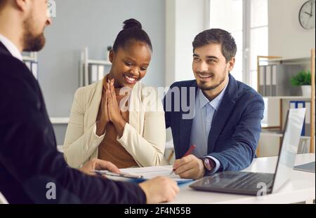 Der glückliche kaukasische Ehemann und seine dunkelhäutige Frau unterschreiben ein Dokument, während sie in einer Bank sitzen. Stockfoto