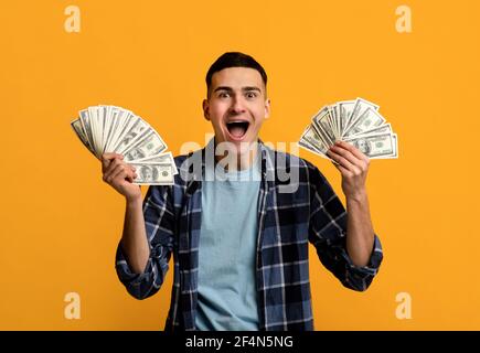 Cool Hipster Kerl hält zwei Fans von Geld, schreien WOW in Aufregung auf orangen Studio-Hintergrund Stockfoto