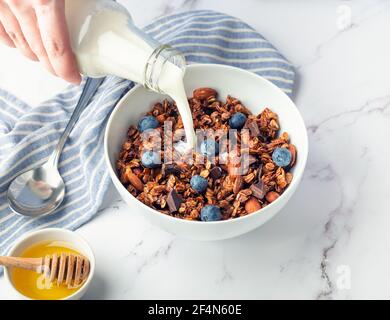 Hausgemachtes Müsli, Müsli mit Stücken von dunkler Schokolade, Nüssen, Heidelbeeren und Milch in Schüssel auf weißem Marmor Hintergrund gießen. Gesundes Frühstück. Stockfoto