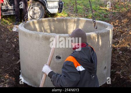 Ein Lader senkt einen Betonring in ein gegrabenes Loch, um einen Klärgruben zu bauen. Ein Arbeiter installiert einen Abwasserkanal in den Boden. Stockfoto