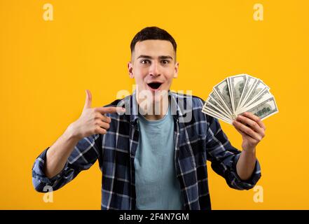 Aufgeregt Millennial Kerl zeigt auf Geld in seiner Hand, zeigt seinen großen Sieg über orange Studio Hintergrund Stockfoto