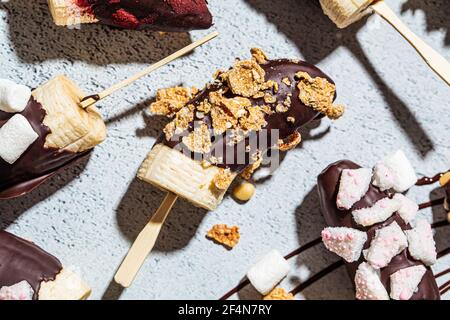 Bananen in dunkler Schokolade mit verschiedenen Belägen auf einem Stock, grauer Hintergrund. Gesundes veganes Dessertkonzept. Stockfoto