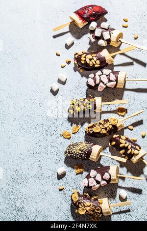 Bananen in dunkler Schokolade mit verschiedenen Belägen auf einem Stock, grauer Hintergrund. Gesundes veganes Dessertkonzept. Stockfoto