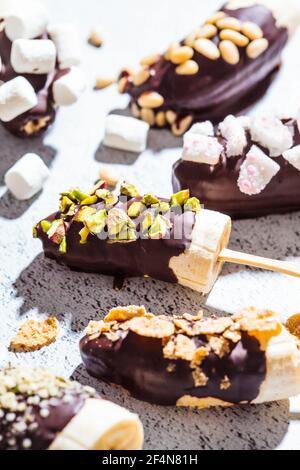 Bananen in dunkler Schokolade mit verschiedenen Belägen auf einem Stock, grauer Hintergrund. Gesundes veganes Dessertkonzept. Stockfoto