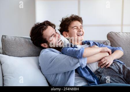 Familienwochenende. Liebevoller Vater kitzelt seinen lachenden Sohn und verbringt Freizeit zu Hause zusammen Stockfoto