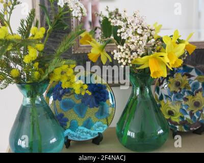 Ideen für eine Frühlingsblumenanzeige auf einem Kaminsims: Zwei kleine farbige Glasvasen mit Frühlingsblumen & verzierten japanischen porzellantellern Stockfoto