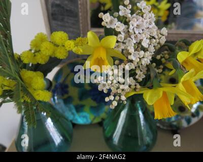 Ideen für eine Frühlingsblumenanzeige auf einem Kaminsims: Zwei kleine farbige Glasvasen mit Frühlingsblumen & verzierten japanischen porzellantellern Stockfoto