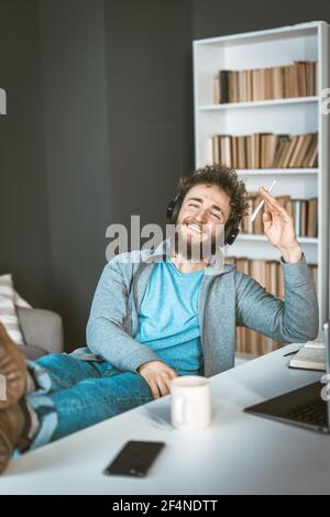 Glücklicher Student zu Hause. Fernlernen des Konzepts. Ein junger Mann mit Kopfhörern sitzt lächelnd vor einem Computer. Stockfoto
