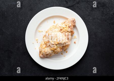 Süßes Mandelcroissant auf weißem Teller, schwarzer Betonhintergrund, Draufsicht. Französisches Gebäck Stockfoto