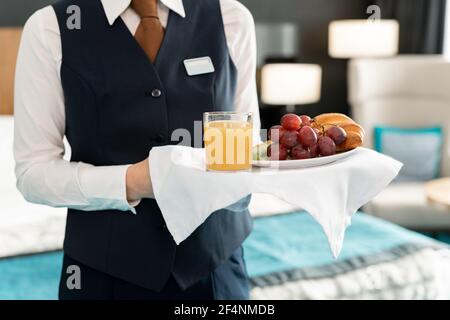 Mittelteil der jungen eleganten Arbeiterin des Hotels, die Tablett hält Mit Frühstück, während man vor der Kamera gegen das Bett steht Stockfoto