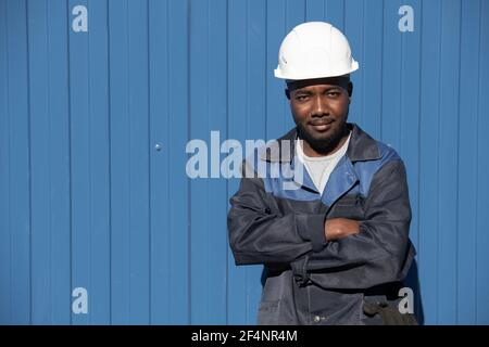 Glückliche junge Fabrikarbeiter der afrikanischen Ethnie in Arbeitskleidung und hardhat kreuzt seine Arme an der Brust, während er davor steht Der Kamera im Freien Stockfoto
