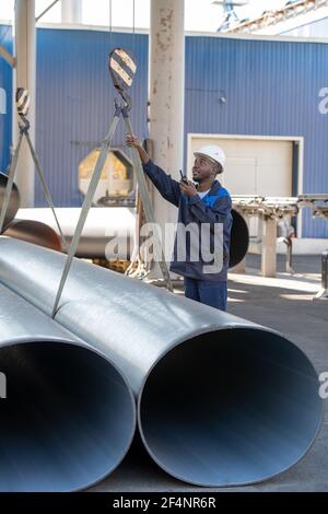 Junge Fabrikarbeiter afrikanischer Ethnie in Arbeitskleidung und Hardhat Mit Walkie-Talkie, während die Kontrolle über das Heben von großen Rohren während Arbeiten Stockfoto