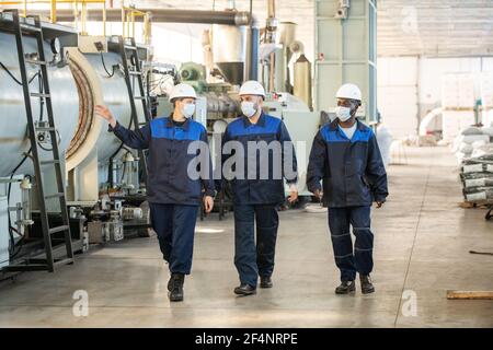 Drei junge interkulturelle Arbeiter der großen zeitgenössischen Fabrik ziehen entlang Industrieanlage und Diskussion neuer Verarbeitungsanlagen Stockfoto