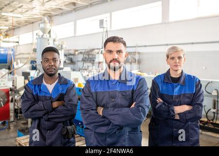 Drei junge interkulturelle Arbeiter der modernen Pflanze in Overalls stehend Vor der Kamera gegen Industriemaschinen in großer Werkstatt Stockfoto