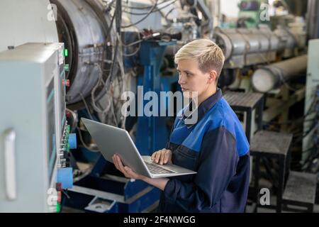 Junge ernsthafte blonde Arbeiterin Overalls mit Laptop während Online-Suche nach technischen Daten gegen Industrieanlagen in der Werkstatt Stockfoto